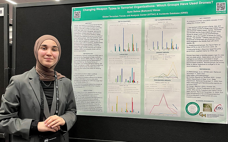 A young woman in a green jacket and a head scarf stands in front of a poster with graphs.
