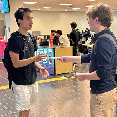 Two men in short pants in a room with a tile floor face each other as one hands the one of the left a piece of paper.