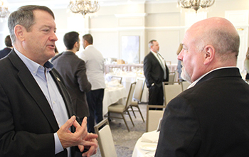 Two men in dark suits stand face to face in a room with chandeliers.