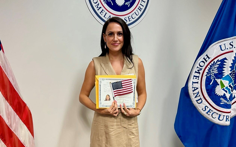 A woman with dark hair stands between flags holding a certificate.
