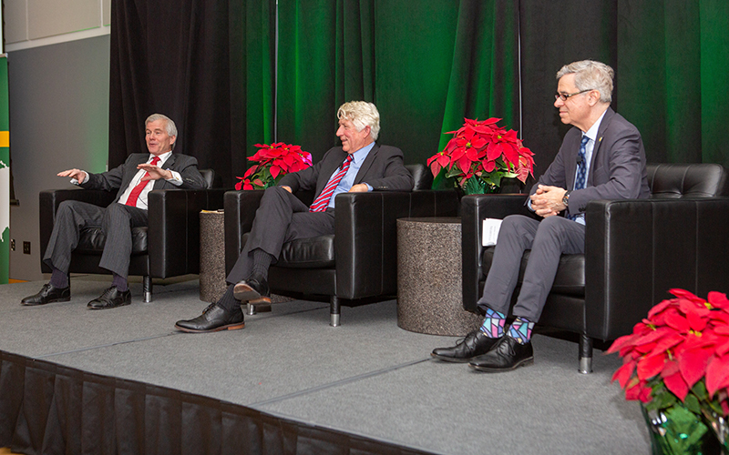 Three men in business suits sit on a stage with potted plants between them.