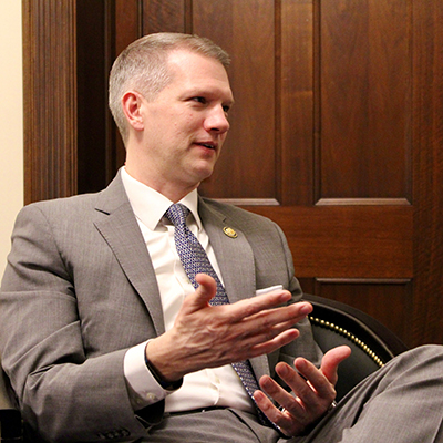 A man with short light hair in a beige suit sits in a chair and gestures with his hands.