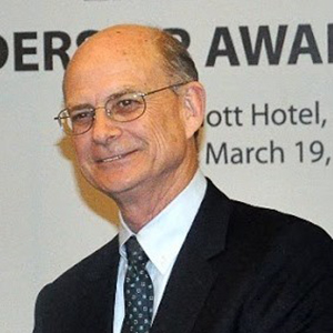 A man in a dark jacket and blue necktie smiles off camera.