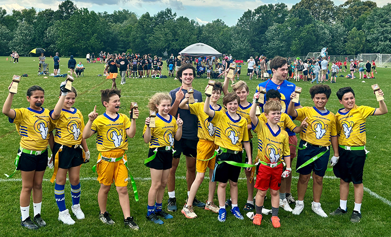 A group of kids wearing yellow flag football shirts stand in front of a man wearing a blue shirt and wave.