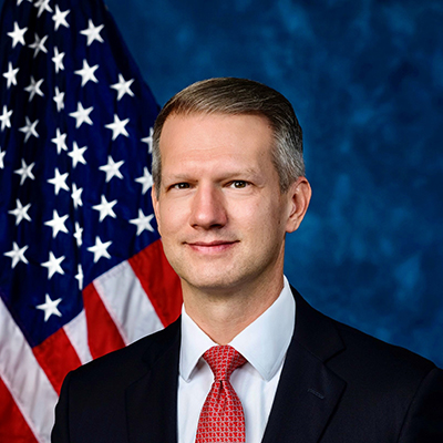 A man in a black suit, white collared shirt, and red tie stands in front of an American flag