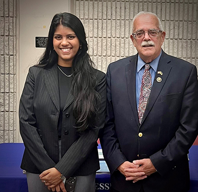 A woman with long dark hair in a dark jacket stands next to a Congressman in a dark suit and tie.
