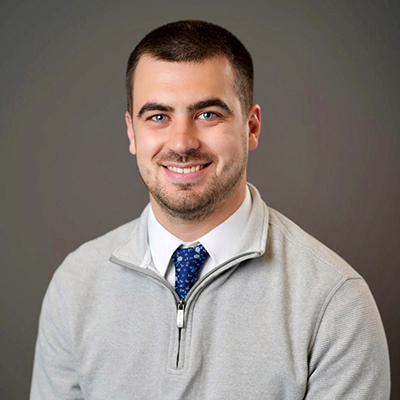 A man in a gray sweater and blue necktie smiles at the camera.