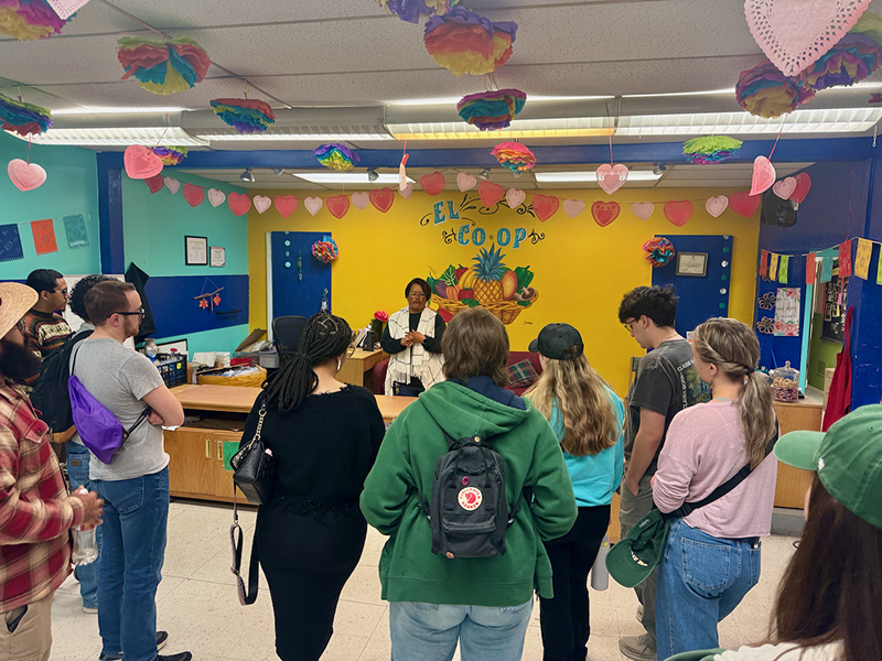 A woman in a black long-sleeved shirt and white and black vest stands in front of a bright yellow wall with the words ‘El Co-op’ and a bowl of fruit painted on it and talks to a group of students.