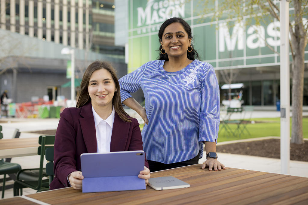 Two female students smiling in front of Van Metre Hall. One is sitting and wearing a maroon jacket and white collared shirt while the other is standing and wearing a blue shirt.