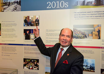 James P. Danoy wearing a dark suit and a red necktie points to a photo of himself with President Obama on a wall.