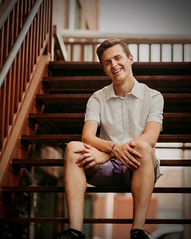 A young man in shorts and short sleeves sits on wooden steps smiling at the camera.