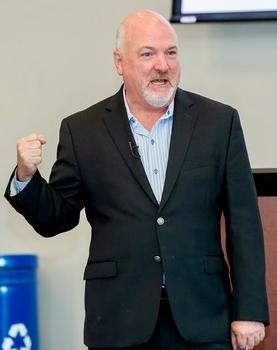 A man in a dark jacket and short white beard makes a fist to make a point.