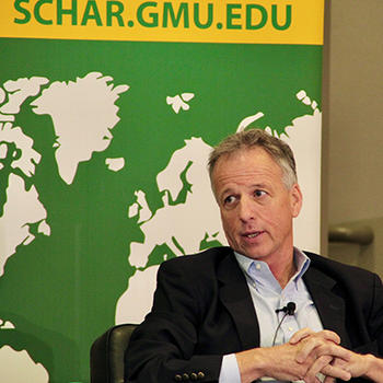 A man in a black jacket and blue shirt with a microphone on his shirt sits in front of a map of the world.