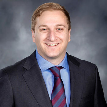 A man with light hair in a dark jacket, blue shirt, and stripped tie smiles at the camera.