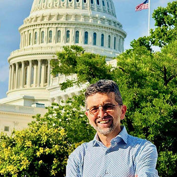A man in a blue shirt and eyeglasses poses in front of the U.S. Capitol building.