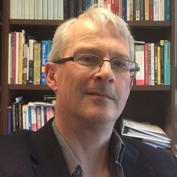 A man wearing glasses, a black jacket, and gray collared shirt stands in front of a book case